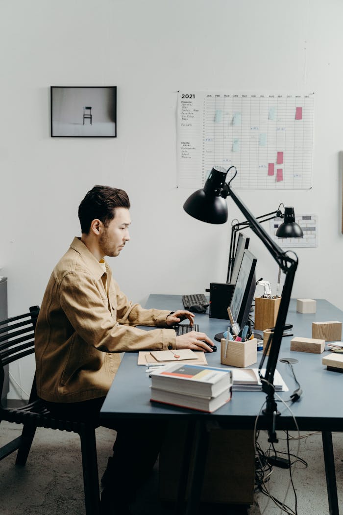 Man in brown jacket typing on a computer at a modern office desk with book stacks and lamps.