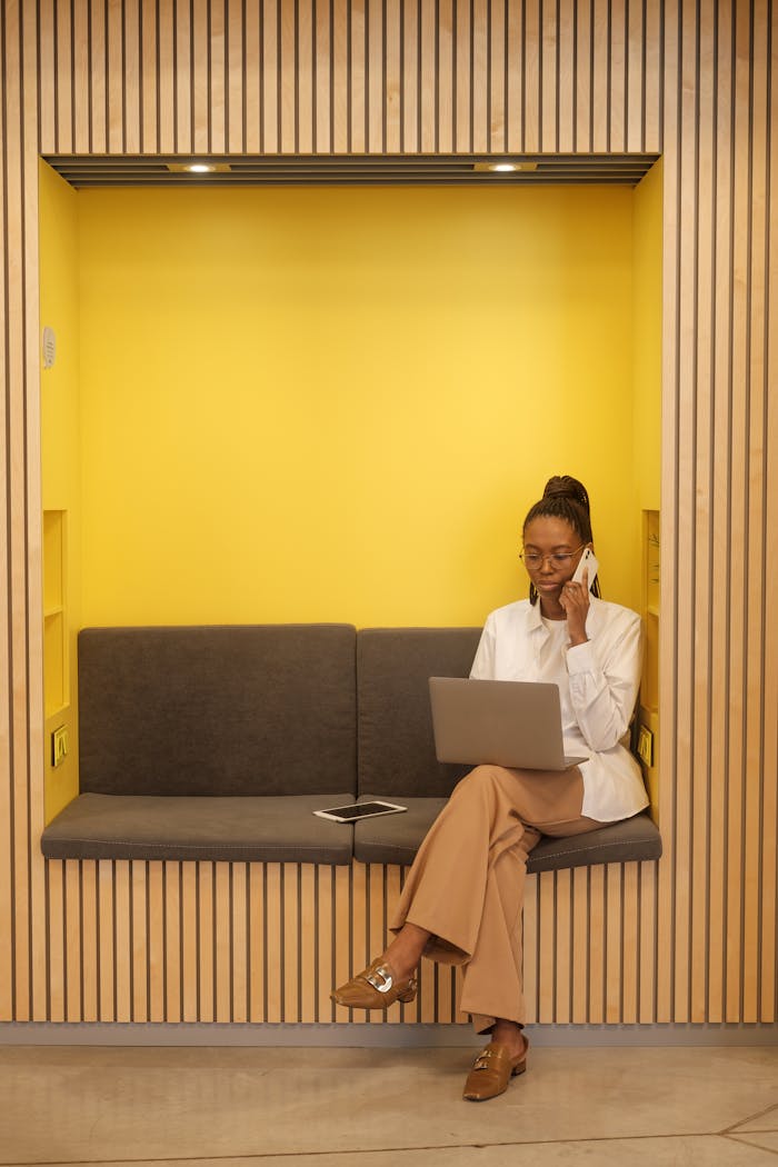 Black woman with braided hair multitasking on phone and laptop in a cozy office nook.